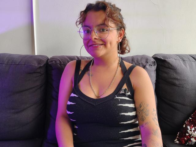 DarcyTear with curly brown hair, glasses, and a smile, wearing a black, zebra-striped tank top, and a chain necklace, sitting on a gray couch with tattoos on her arm.