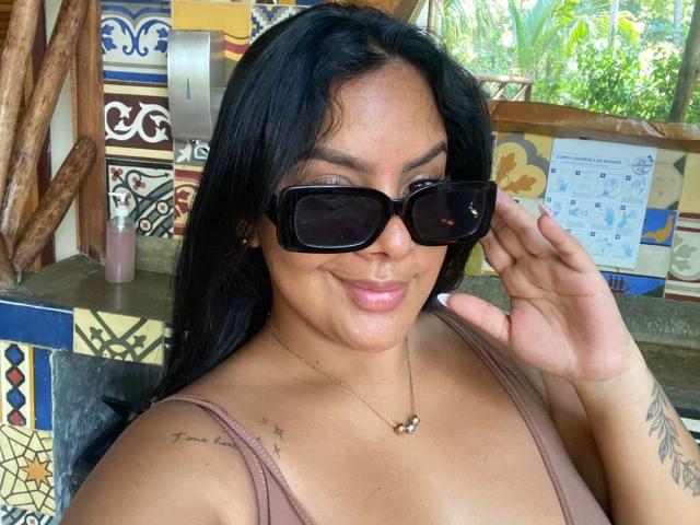 AmarantaCarey with black hair, brown skin, wearing black sunglasses, a brown tank top, and a gold necklace, holding her sunglasses, showing a tattoo on her right shoulder.