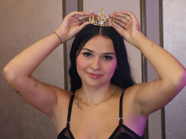 AlahiaStorm with long black hair, wearing a gold crown, black sheer bra, smiling, arms raised, showing tattoos and a necklace, pinkish light on right side.