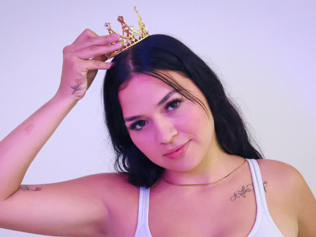 AlahiaStorm, con i capelli neri, indossa una canotta bianca, ha una corona d'oro sulla testa, dei tatuaggi sul braccio e sul petto, e un viso carino e sensuale..