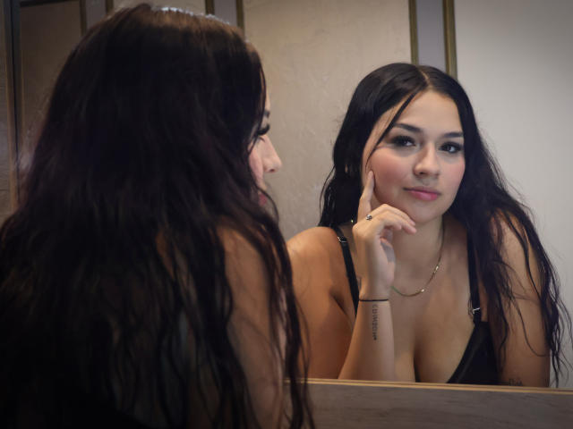 AlahiaStorm with long dark hair, small nose ring, black tank top, looking at mirror, gently touching cheek, smiling slightly, showing small tits, subtle makeup, gold necklace, bracelet.