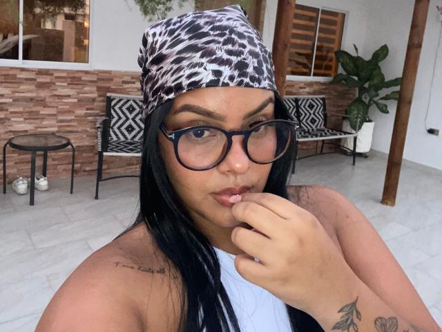 AmarantaCarey with long black hair, glasses, leopard print beanie, white tank top, tattoos on arm and chest, finger in mouth, looking at camera, standing outside.