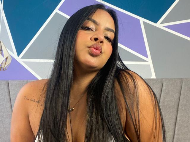 AmarantaCarey with long black hair, pouty lips, and a tattoo on her right shoulder, wearing a necklace, looking at the camera. Boobs visible, grey background.