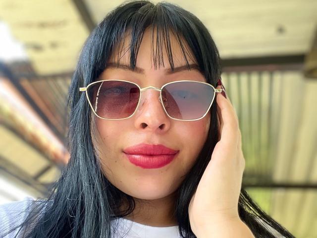 KateWelsin with black hair, pink lipstick, and gold-rimmed sunglasses, touching her ear with a hand, wearing a white shirt, smiling subtly.