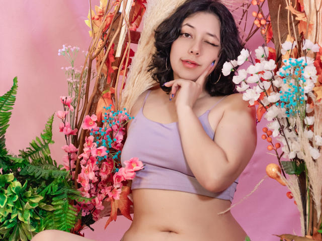 MillieRoy with wavy black hair, winking, in a lilac crop top, showing her tits, surrounded by colorful flowers and greenery, touching her cheek with a playful expression.