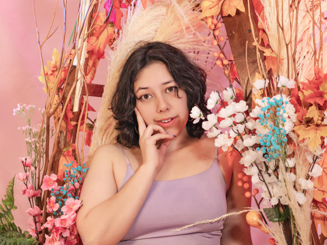 MillieRoy with curly black hair, purple tank top, touching her cheek, surrounded by colorful flowers and foliage. She's looking at the camera with a cute, relaxed expression.