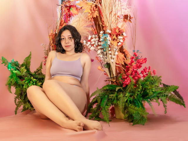 MillieRoy with curly dark hair, wearing a lilac sports bra and beige panties, sitting with legs crossed, surrounded by green ferns, pink flowers, and red foliage.