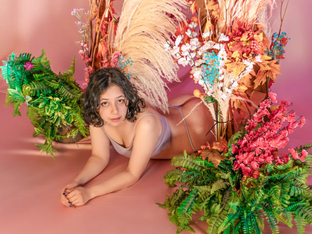 MillieRoy lying on her stomach, wearing a grey bra and thong, with curly dark hair, surrounded by green ferns, pink flowers, and puffy beige grasses on a pink surface.