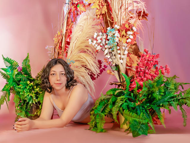 MillieRoy with curly brown hair, light skin, and small tits, wearing a white tank top, lying on pink floor with green ferns and colorful floral arrangements in the background.