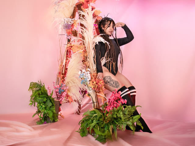 BeckyHuston with long braids, black long-sleeve top, and thigh-high socks, standing beside floral arrangements, showing a tattoo on her right thigh, against a pink backdrop.