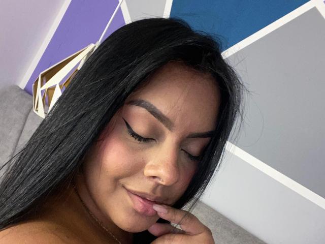 AmarantaCarey has long black hair, closed eyes with winged eyeliner, and a subtle smile, gently touching her pink lips with a finger. She's wearing a gold necklace.