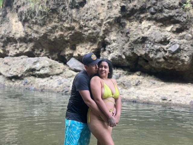 LianaAndAsaff in a yellow bikini, standing in shallow water, with a man in a black cap and blue shorts kissing her neck, his hand on her ass.