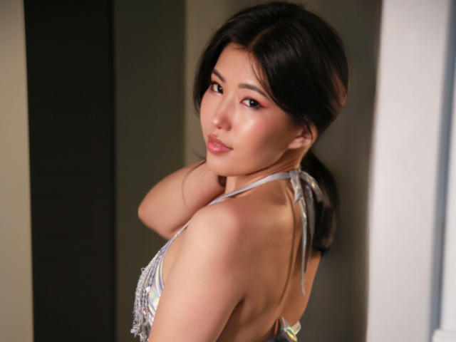 WendyFire with dark hair, looking over her shoulder, wearing a silver halter top, showing her bare back and ass. Her hand touches her neck, with soft, confident eyes.