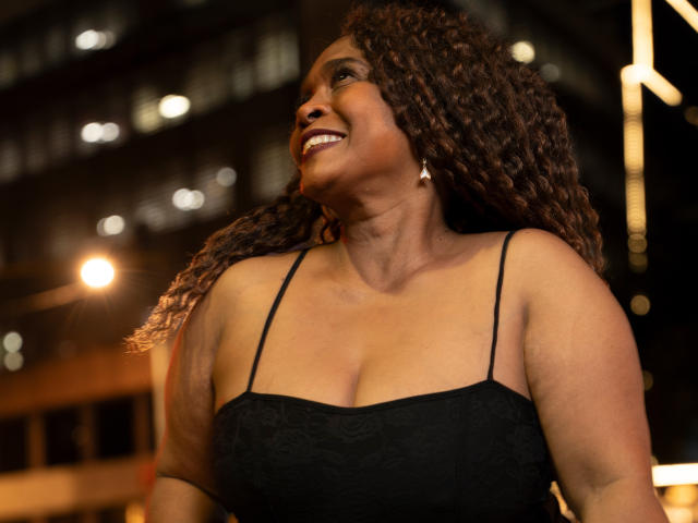 AinaEbonyQueen, une femme noire aux formes généreuses, avec des cheveux bruns bouclés, souriante, portant une robe noire à bretelles fines, montrant ses seins, debout la nuit avec des lumières de rue chaudes..