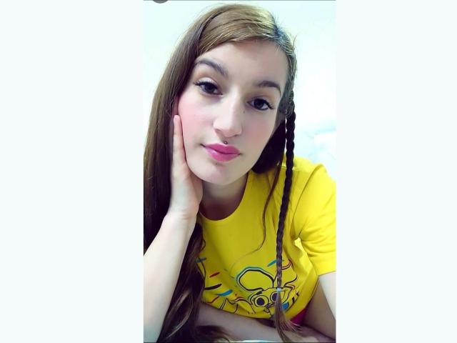 RitaJonness has long brown hair, a yellow shirt with a colorful graphic, and pink lipstick, resting her cheek on her hand with a slightly pouty expression.