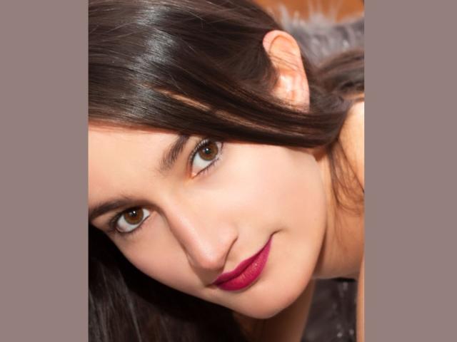 RitaJonness has brown eyes, dark straight hair, and pink lipstick, smiling with a slight tilt of her head. Her fair skin is smooth and she's looking directly at the camera.