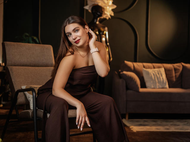 MilanaKitty with long brown hair, wearing a strapless brown dress, sitting on a chair, hand on her cheek, smiling, showing her tits, in a dimly lit room.