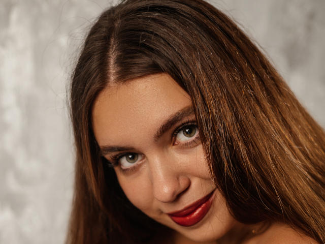 MilanaKitty has brown hair, green eyes, and red lipstick. Her slightly upturned nose and subtle smile are visible. Her tits aren't shown, just her face.