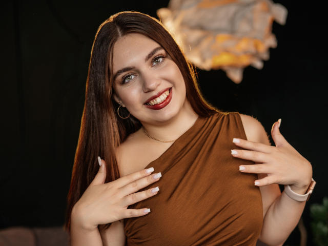 MilanaKitty with long brown hair, smiling, wearing a brown one-shoulder top, hands on her tits, red lipstick, hoop earrings, white bracelet, and nail polish in a dark room.