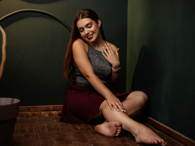 MilanaKitty with long brown hair, gray sleeveless top, red skirt, sitting on wooden floor, smiling, touching her chest, bare feet, green wall, dark brown lamp in the background.