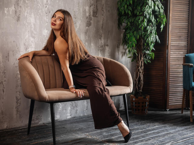 MilanaKitty with long brown hair, wearing a brown dress, leaning on a beige chair, black heels, looking back, showing her ass, in a room with a potted plant.