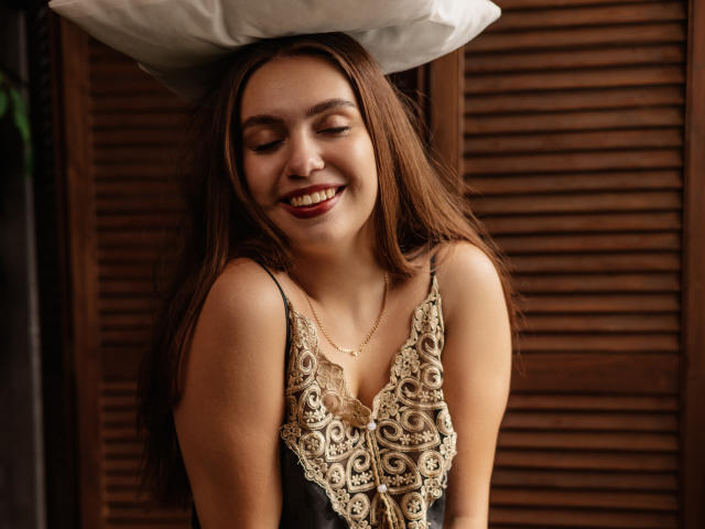 MilanaKitty with long brown hair, smiling with eyes closed, wearing a lace-trimmed black camisole, white pillow above head, brown wooden background, and a gold necklace.