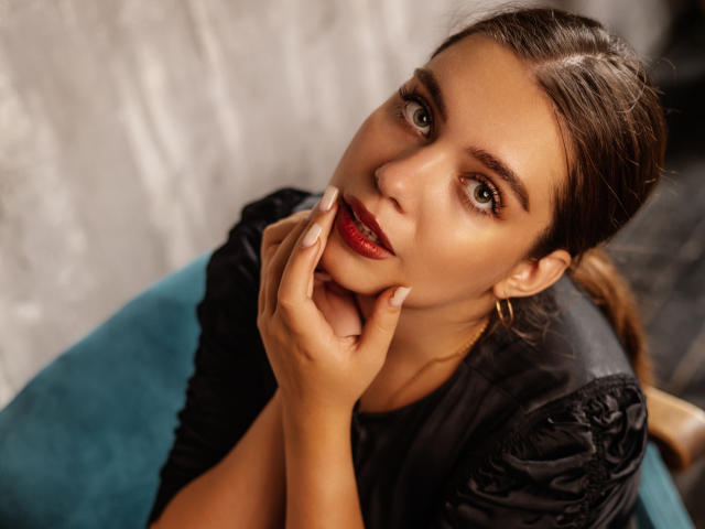 MilanaKitty with brown hair, green eyes, and red lipstick, holding her chin with manicured fingers, wearing a black top, gold earrings, and a blue seat.