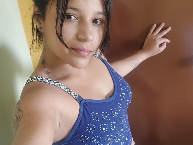 LorenaHt with dark hair, blue sparkly tank top, small tits, tattoos on shoulders, right arm raised, brown door in background, slight smile, brown eyes, light skin.