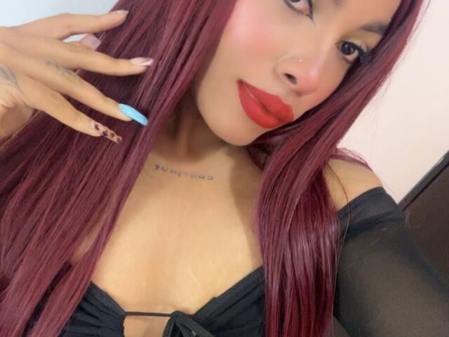 LexieBloom with red lipstick, long purple hair, black off-shoulder top, blue and brown nails, tattoo on chest, and a cute nose ring, showing off her tits.