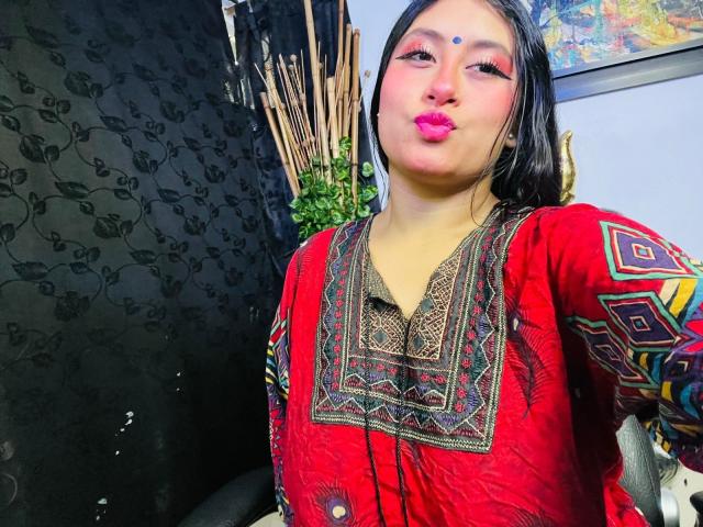 KalpanaAisha with puffy pink lipstick, dark straight hair, red traditional dress with intricate patterns, and a bindi on her forehead, making a pouty face. Boob outline visible.