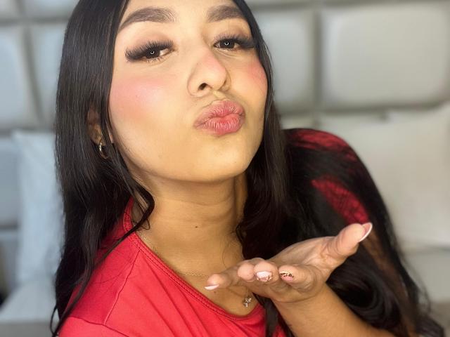 SofiaJames with long black hair, red shirt, and pink lipstick, pouting her plump, shiny ass-like lips, holding up her manicured finger with a gold ring.
