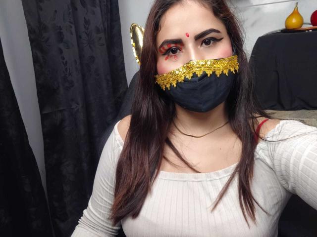 DaliaDubey with dark hair, wearing a black mask with gold lace, red makeup on her cheeks, white ribbed shirt, red strap, and a red dot on her forehead.
