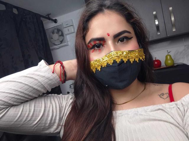Dalia Dubey, met lang bruin haar, draagt een zwart masker met gouden rand, rode oogmake-up, een wit geribbeld topje, een rode beha-band, rode armbanden en een kleine rode stip op haar voorhoofd..