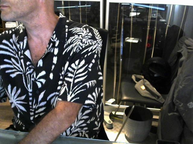 Oliviermodeltest lleva una camisa con un estampado de hojas en blanco y negro, mostrando un cuello y parte superior del pecho de piel clara y sin vello, sentada en una silla con zapatos negros cerca..