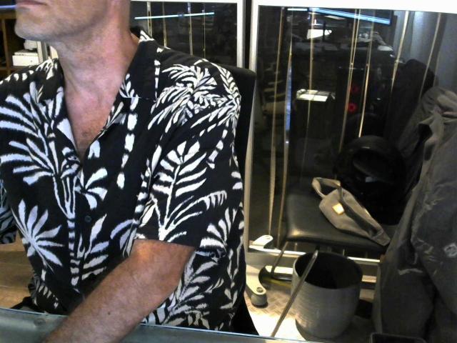 oliviermodeltest portant une chemise avec un motif de feuilles noir et blanc, visible du cou à la poitrine, assise dans un fauteuil de coiffeur, la tête hors du cadre, les mains reposant sur un comptoir..
