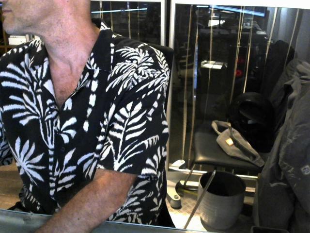 Modelo Olivia, con una camisa de color blanco y negro con estampado de hojas, mostrando un cuello y parte superior del pecho delgados, de pie junto a una silla con un bolso negro y zapatos color beige..