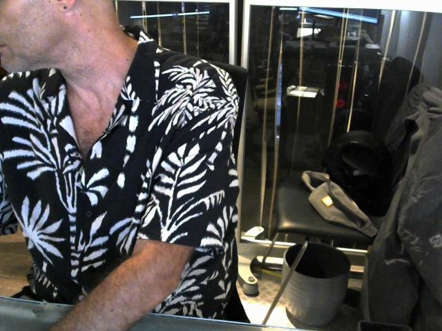Modelo Olivi con la cabeza calva, vistiendo una camisa de color negro y blanco con estampado floral, inclinándose ligeramente hacia adelante. En el fondo, se ven sandalias beige y una chaqueta gris..