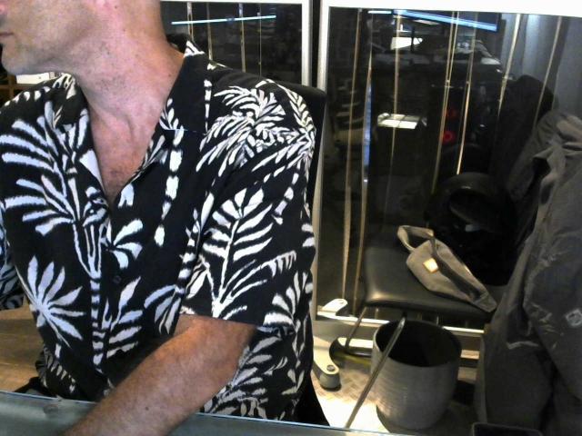 Modelo Olivi, con la cabeza calva, vistiendo una camisa con un estampado de hojas en blanco y negro, tiene un cuello ligeramente musculoso y está de pie en una habitación con una silla y zapatos..