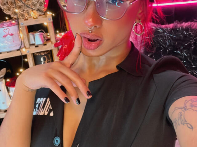 AlexaFoster with red hair, glasses, nose ring, and black shirt, finger near mouth, black nails, tattoo on arm, pink neon light in background.
