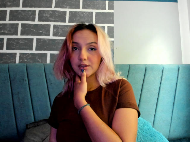 AlterEgoSecond has blonde hair, brown t-shirt, blue nails, finger on chin, teal cushioned bench, gray brick wall, blue wall, light in background, bracelet on wrist, serious expression.