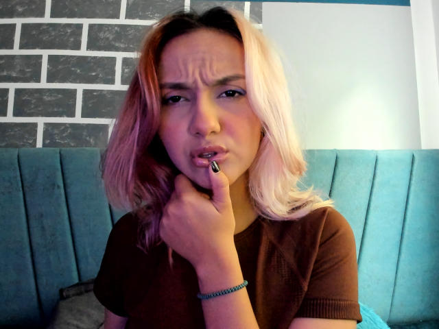 AlterEgoSecond with pink and blonde hair, brown shirt, blue cushioned seat, thinking face, finger on chin, black nail polish, blue bracelet, looking slightly annoyed.