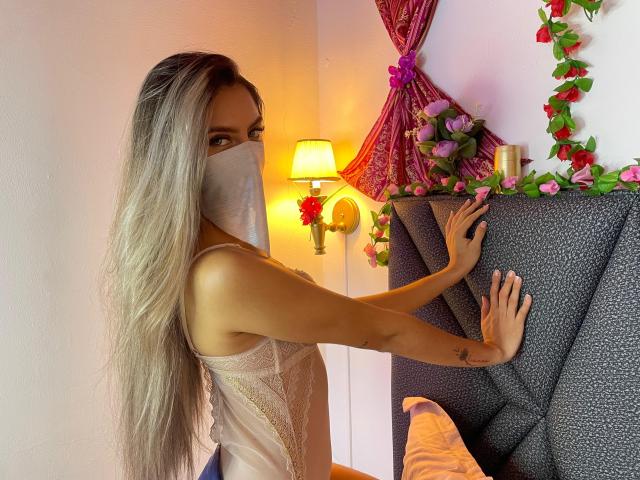 DalilaAliX with long blonde hair, wearing a sheer, lace-up top, silver mask, touching a gray, textured headboard, her hand showing a tattoo, in a warmly lit room.