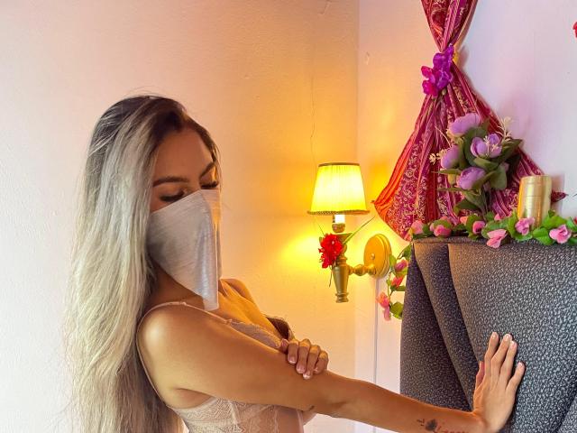 DalilaAliX with long blonde hair, wearing a face mask, and a lace top, arms crossed, standing next to a gray headboard with pink flowers and a lit lamp.