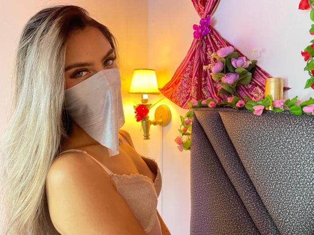 DalilaAliX with long blonde hair, wearing a white face mask and lace top, showing her tits, standing by a lit lamp, pink and purple flowers, and a gray curtain.