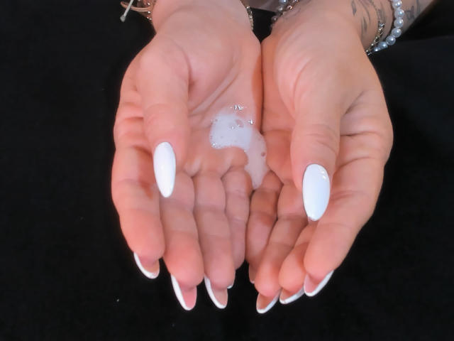 ChristineBray's hands with white nails, holding a small white liquid drop between them, silver bracelet, tattoo visible on her wrist, black background.