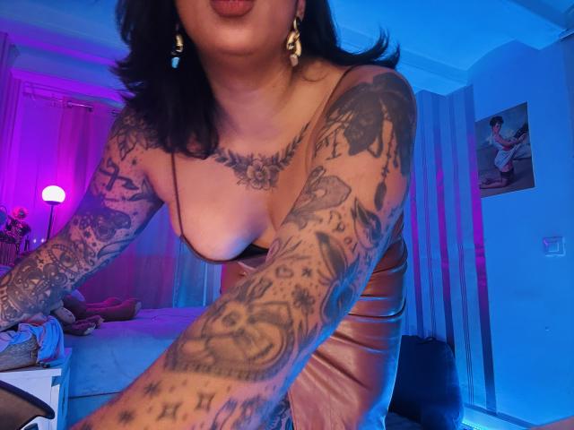 Chili, topless, braccia tatuate pesanti, seni grandi, illuminazione viola e blu, camera da letto, lingua fuori, abito di pelle marrone, orecchini in oro.