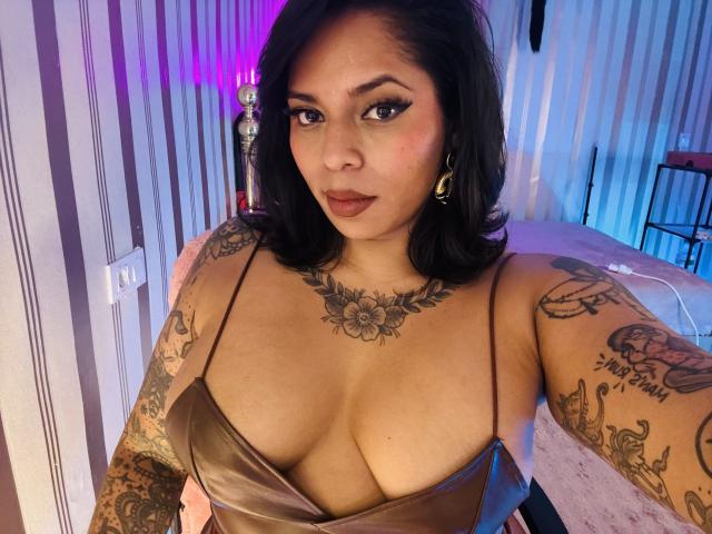 Chili with tattoos, brown dress, cleavage, black hair, looking at camera, pink and blue lights in background, confident expression.