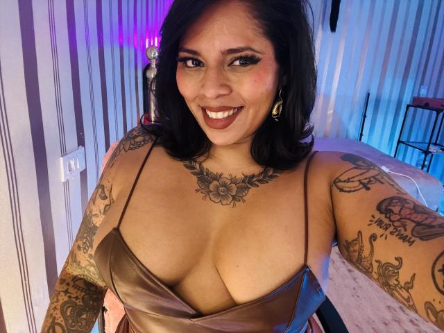 Chili tem seios médios, braços tatuados e um sorriso largo e sexy, vestindo um vestido marrom sedoso e decotado.