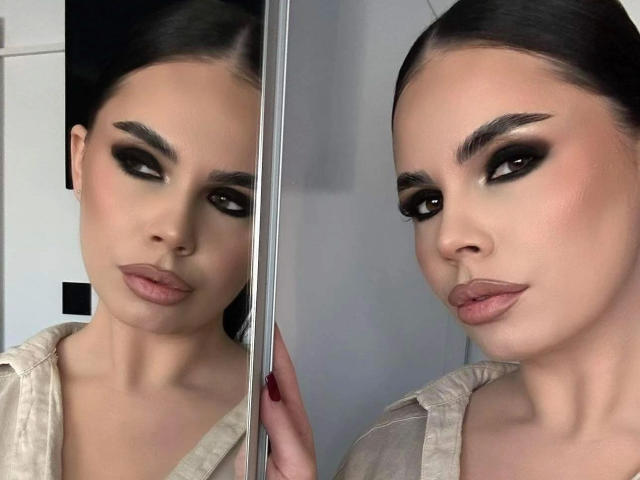 JessicaJolie-hot has dark makeup, full eyebrows, and pink lips. Her reflection shows her holding a mirror, with a beige shirt on. Her pussy is not visible.