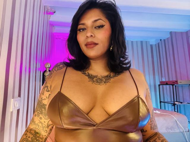 Chili, curvy Latina with big breasts, wearing a metallic bronze bra, tattoos on arms, black hair, confident look, bedroom background.